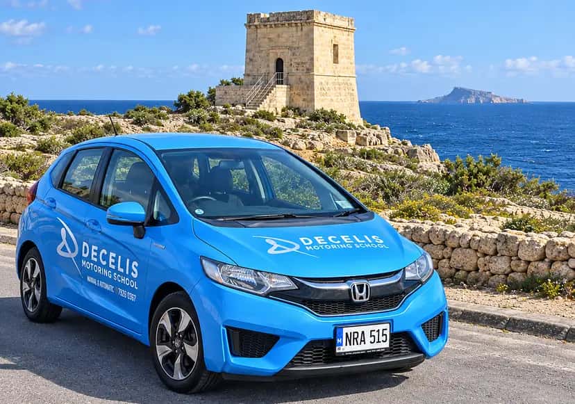 Decelis Motoring School's blue Honda Jazz, plate NRA 515, parked below the Xutu Tower at Wied iż-Żurrieq with Filfla on the sea